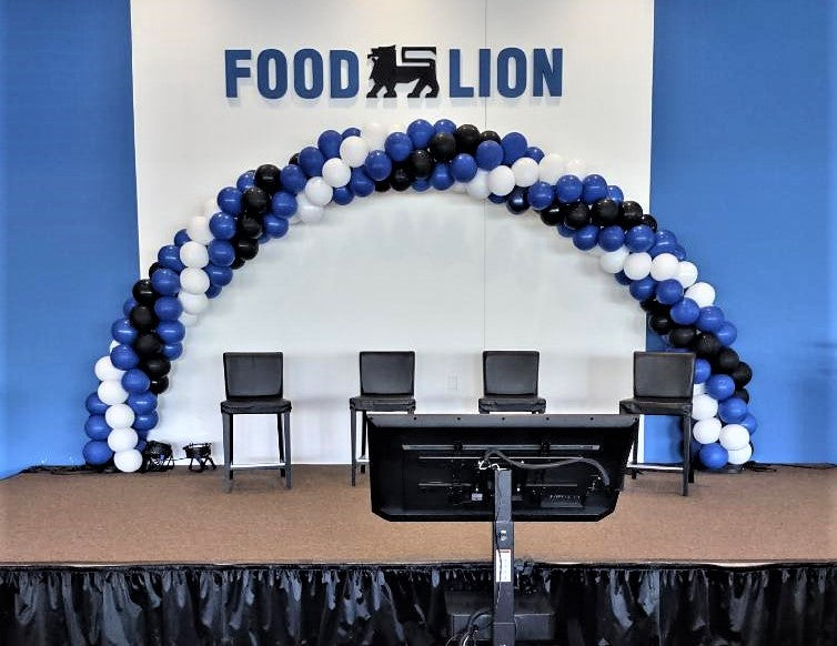 large blue, black, and white balloon arch on stage for Food Lion grocery store with 4 chairs under it.