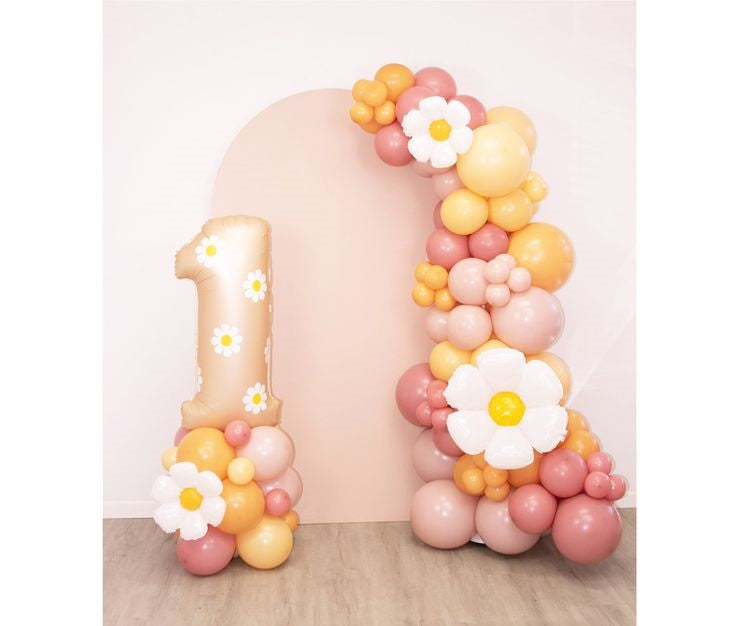 An balloon garland with backdrop foil flowers displayed as a decoration, featuring a large balloon in the shape of a number 1, adorned with balloons shaped like flowers and a crown, in shades of pink, white, and yellow.