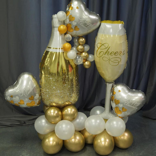 A balloon bouquet featuring a champagne bottle and glass balloons with and bubbles balloons.   anniversary and heart balloons with 'CHEERS' and 'HAPPY ANNIVERSARY' on it, along with other round and foil balloons in gold, silver, and white colors