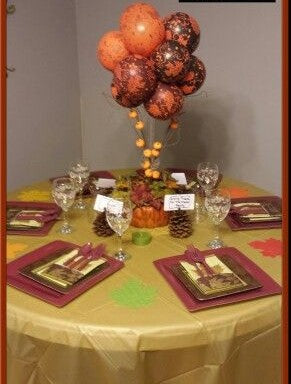 A fall-themed topiary centerpiece featuring a tree with orange, brown, and gold balloons, as well as red and gold leaves and flower arrangements on a golden tablecloth.