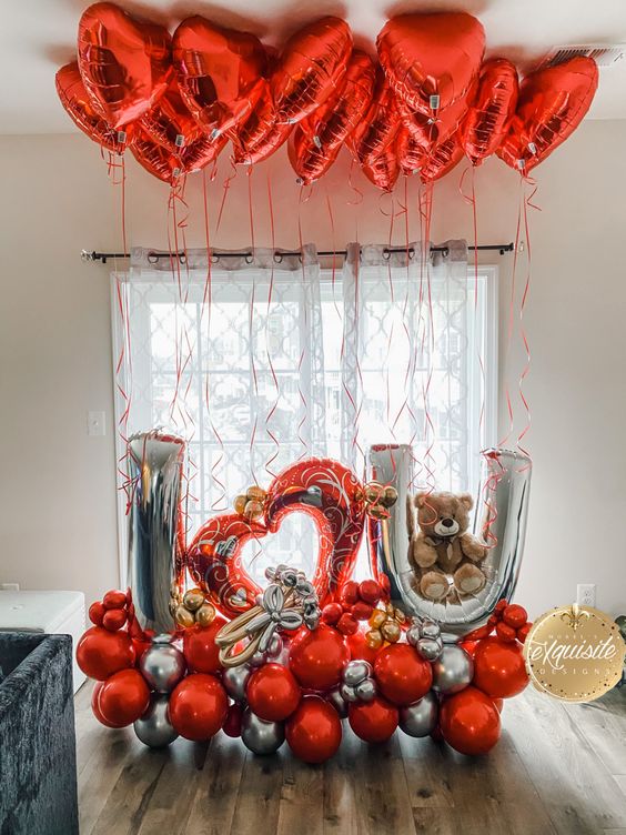 A Valentine's Day themed balloon bouquet featuring red and silver balloons with the phrase 'I (heart) U' along with a stuffed teddy bear and sculpted balloon flowers, set against a backdrop with a window and curtains and heart balloons on ceiling.