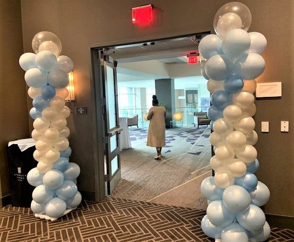 Two balloon columns made of white and blue balloons, with a clear topper on top, standing in a room with a person in the background.
