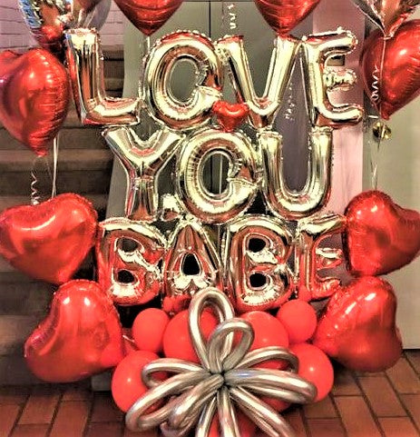 A valentine's day balloon bouquet with gold balloon letters that spell  "love you babe" surrounded by red foil and mylar red heart shaped balloons and red round balloons and gold flower balloons