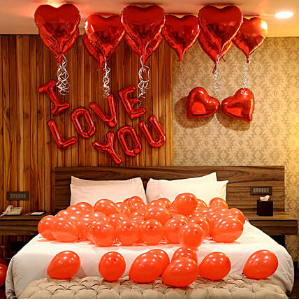 A room with a bed and Valentine's Day decorations, including red balloons on the bed, red foil heart-shaped balloons on the ceiling and wall  and the phrase 'I love you' written with red letter balloons and red heart shaped balloons in the ceiling and wall.