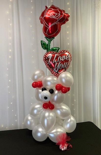 A sculptured balloon teddy bear in white and red with a foil heart balloon that says "I Love You" and a red rose, accented with mylar balloons and chocolate kisses.