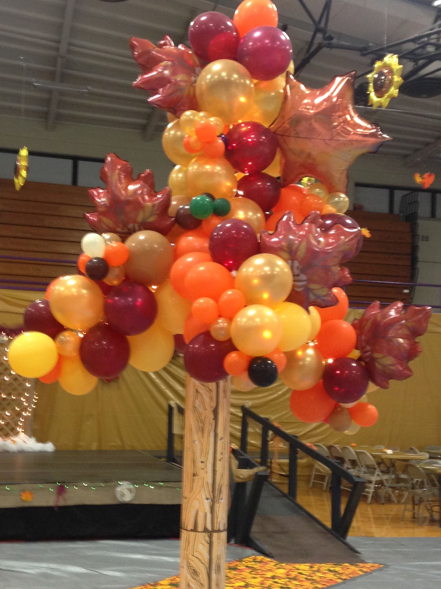 A balloon tree sculpture with a wooden stump, decorated with a variety of fall-colored balloons in shades of orange, red, yellow, brown, and burgundy, set against a backdrop of a Thanksgiving-themed event.